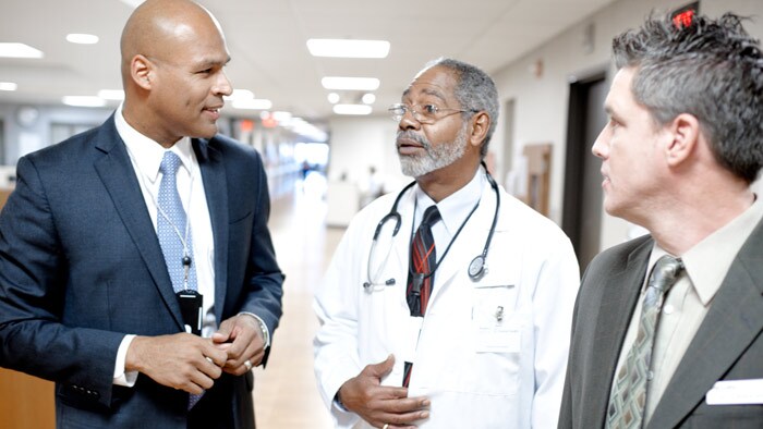 A doctor and a man in suit walk through a hallway as another person in suit looks at both